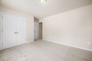An empty room with beige walls, beige carpet, a ceiling light, a closed double-door closet, and an open door leading to another area.