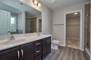 A modern bathroom with a double vanity, large mirror, toilet, walk-in shower, towel rod, and a door leading to a walk-in closet. The floor has wood-like tiles, and the room is lit by ceiling lights.