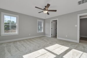Empty, well-lit bedroom with gray walls, beige carpet, two windows, a ceiling fan, and an open door leading to an adjacent room.