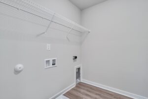 Empty laundry room with light gray walls, wire shelf, and utility hookups for a washer and dryer. Wood-look flooring and a small alcove for venting or additional hookups.