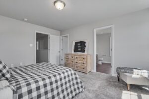 A bedroom with a gray checked bedspread, a wooden dresser with a chalkboard, and an upholstered bench. An open door reveals a bathroom. Walls are light gray, and the carpet is gray.