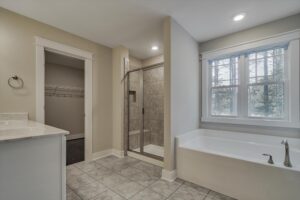 A bathroom with tiled floors, a bathtub, a glass-enclosed shower, a window, a countertop with a sink, and a walk-in closet.
