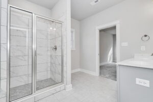 Modern bathroom with marble tiles, glass shower door, white vanity, and a doorway leading to another room.