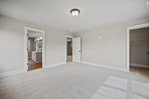 An empty bedroom with beige walls and carpet, white trim, a closed door, an open door leading to a bathroom, and a closet with a hanging rod.