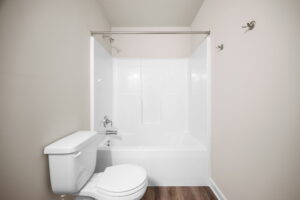 A bathroom with a white bathtub-shower combo, a white toilet, and beige walls with wooden flooring. Two towel hooks are visible on the right wall.
