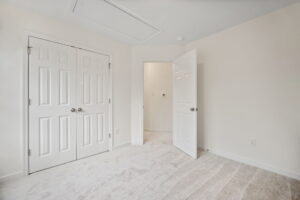 A small, empty room with beige carpet, white walls, a closed double-door closet, and an open door leading to a hallway.