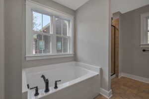 Modern bathroom featuring a large soaking tub with black fixtures, a window view, and a shower with beige tiles in the background.