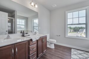 A bright bathroom featuring a double sink vanity with wooden cabinets, a toilet, a large window, and a bath mat on the floor.