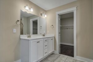 Bathroom with a double sink vanity, wall-mounted mirrors, overhead lighting, and white cabinetry. There's a walk-in closet visible through an open door on the right. The tile floor is gray.