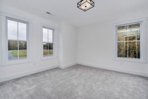 Empty room with white walls and carpeted floor, featuring two large windows on the left wall and one on the right, allowing natural light to enter. A modern light fixture hangs from the ceiling.