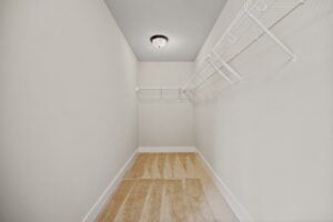 An empty, narrow walk-in closet with beige carpet, white walls, a ceiling light, and metal wire shelving on both sides.