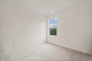 Small, empty room with light-colored carpet. Walls are painted white. A single window provides natural light and a view of greenery outside.