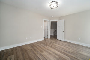 An empty, light-filled room with beige walls, wood flooring, a modern ceiling light fixture, two open doors, and white trim doors and baseboards.