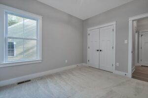 A simple, empty room with gray walls, beige carpet, a window, and double-door closet. There are open doors to the hallway visible on the right.