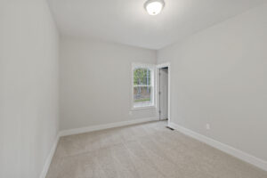 Empty room with beige carpet, gray walls, a window, and a closed door on the right side. Ceiling light fixture is on.