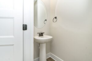 A small bathroom with a white pedestal sink, round mirror, and an open door on the left.