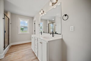 Modern bathroom with large mirror, double sink vanity with white cabinets, black fixtures, and a walk-in shower. Natural light enters through the window. Honey-colored wood flooring.