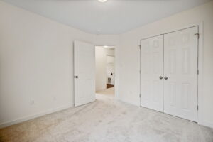 Empty room with light-colored carpet, white walls, and two white doors. One door is open, revealing an additional room with a closet space. Ceiling light fixture is visible.