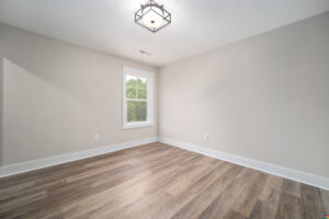 An empty room with light-colored walls, a window with white trim allowing natural light, and wood-style flooring. A rectangular light fixture is mounted on the ceiling.