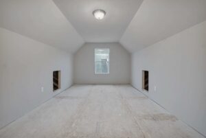 An unfinished attic room with slanted walls, a ceiling light, a central window, and two small alcoves on either side. The floor is bare, and the walls are painted white.