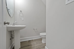A small bathroom with a pedestal sink, round mirror, towel ring, toilet, and white walls. The floor has light wood-like flooring, and there is a wall vent near the baseboard.
