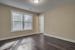 A room with beige walls, dark wooden flooring, a ceiling light, and a window with white trim. An open door is visible in the right corner.