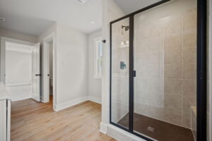 A modern bathroom featuring a walk-in shower with a glass door, beige tiles, and a separate toilet area with a white door. The floor is made of light-colored wood.