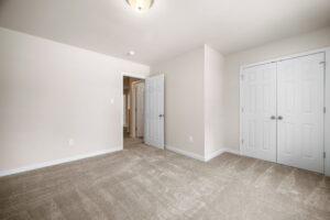 A small room with beige carpet, off-white walls, a ceiling light, double door closet, and an open doorway showing a hallway.