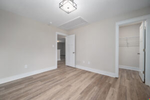An empty room with light gray walls, wood flooring, a white door, a closet with shelves, and a modern ceiling light fixture.
