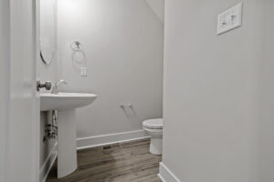 A small bathroom with a pedestal sink, wall-mounted faucet, circular mirror, toilet, and light grey walls with wood-look floor tiles.