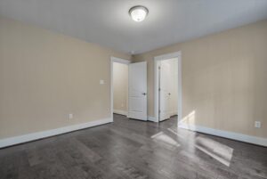 Empty room with beige walls, dark hardwood floors, a ceiling light, and two white doors. One door is open, revealing a lit interior space with white walls.