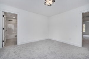 A white room with gray carpet, minimal furniture, and two doorways; one leading to a bathroom and the other to a hallway or another room.