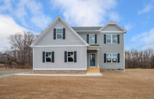 A newly constructed two-story house with gray siding, black shutters, a dark green front door, and multiple windows, surrounded by an unfinished yard with dirt ground, under a partly cloudy sky.
