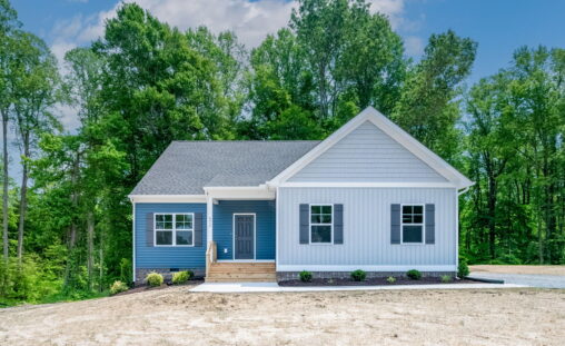 New single-story house with gray siding and a blue front door, surrounded by lush green trees under a clear sky.