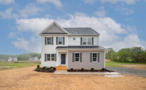Two-story house with a combination of white and gray siding, a porch with steps leading to a blue front door, and small shrubs in front. The background shows open land and a cloudy sky.