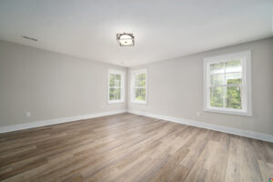 An empty, light-filled room with wooden flooring, beige walls, a square ceiling light, and three windows displaying greenery outside.