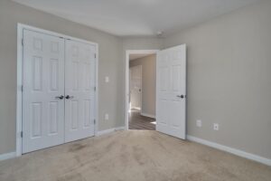 A beige carpeted room with two white closet doors on the left and a white open door on the right leading to a hallway with wooden floors. Walls are painted light gray.