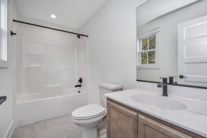 Clean, modern bathroom with a white bathtub, toilet, and sink. The room features a large mirror, light gray flooring, and a small window near the tub.