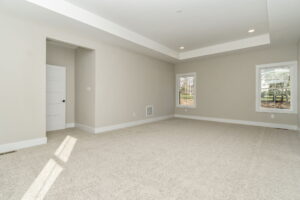 Empty living room with beige carpet, a white door, large windows with a view of trees, and recessed ceiling lights.