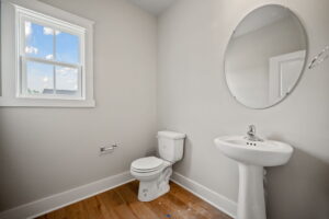 A small bathroom features a round mirror, pedestal sink, toilet, and white walls with a window providing natural light against a wooden floor.