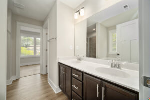 A modern bathroom featuring a double sink vanity with dark cabinets, a large mirror, bright lighting, and an adjacent room visible in the background with a large window.