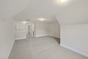 An empty room with beige carpet, white walls, and a slanted ceiling. There are two ceiling lights, a doorway, and a small partition wall in the background.