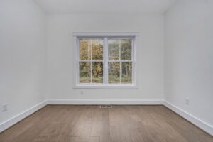 A small, empty room with white walls, hardwood floor, and a central window showing a view of trees outside.