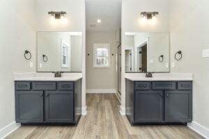 Symmetrical bathroom with two separate gray vanities, each topped with white counters and framed mirrors, under sconce lighting.