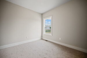 A small, empty room with beige carpet and light gray walls. A single window on one wall lets in natural light, revealing a suburban view outside.