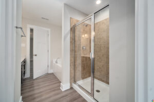 A modern bathroom features a glass-enclosed shower with brown tiles, a white bathtub, a countertop with drawers, and wood-patterned flooring.