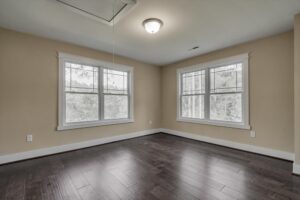 A vacant room with two large windows, beige walls, dark hardwood flooring, and a ceiling light fixture.