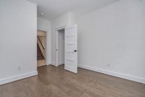 Empty room with light gray walls, wooden flooring, and a white door slightly ajar. An open doorway leads to a staircase. A dark handle is visible on the door, and electrical outlets are on the walls.