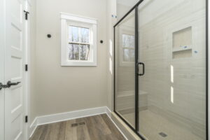 A modern bathroom featuring a glass shower enclosure, beige tiles, a small window, and wooden flooring.