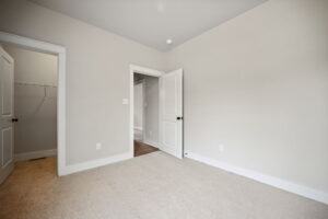 An empty carpeted room with beige walls, a door leading to a walk-in closet on the left, a partially open door in the center, and a small portion of a hallway visible.